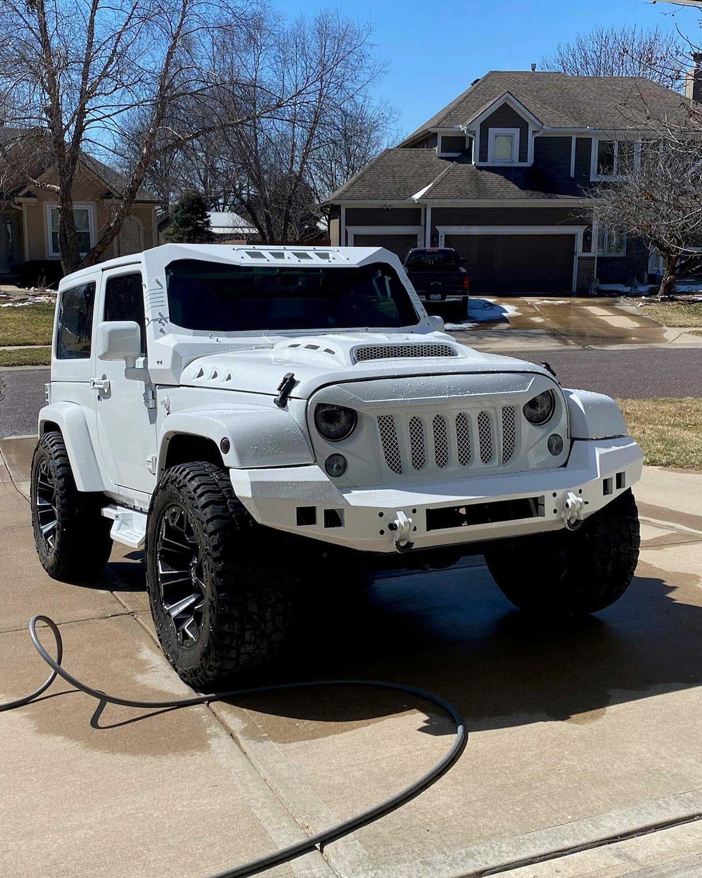 White Jeep Wrangler with black wheels, modified grill, and front bumper, parked on a paved driveway in front of a house.