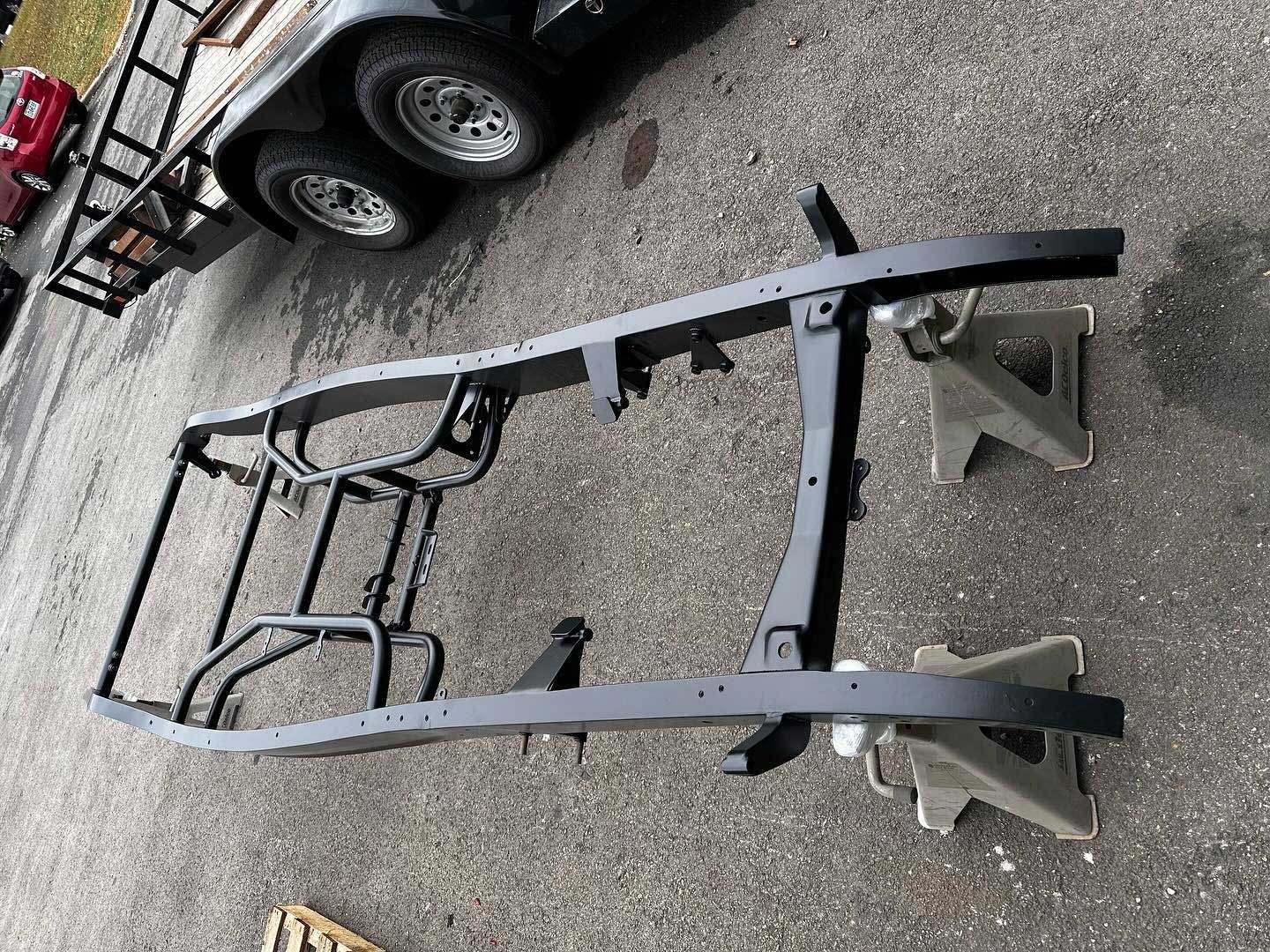 Black vehicle frame on jack stands, parked outdoors, near a trailer.