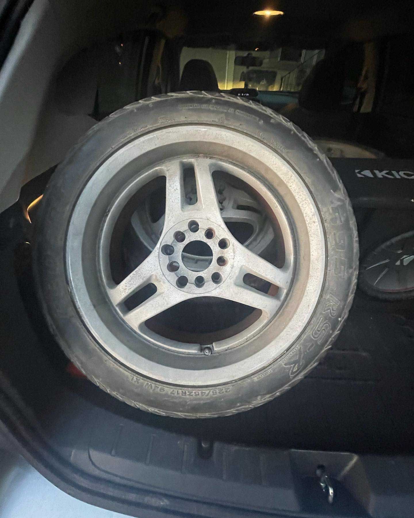 Silver car wheel with a tire in the trunk of a vehicle, next to a black speaker.