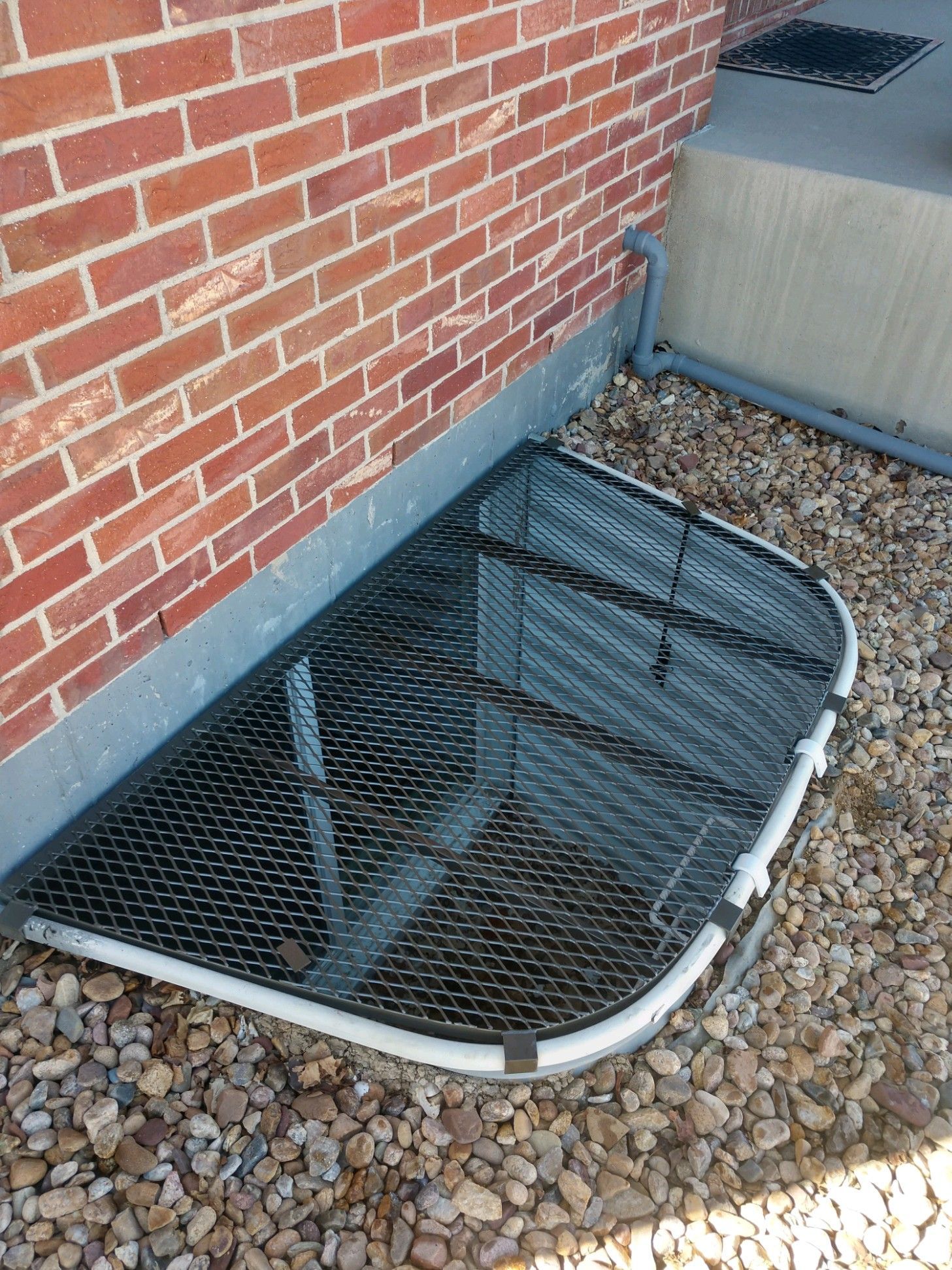 A window well with a screen on it is next to a brick wall.