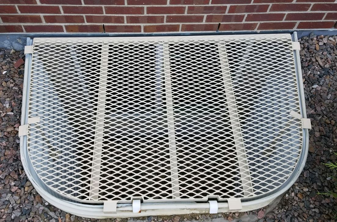 A window well with a metal grate on it is sitting next to a brick wall.