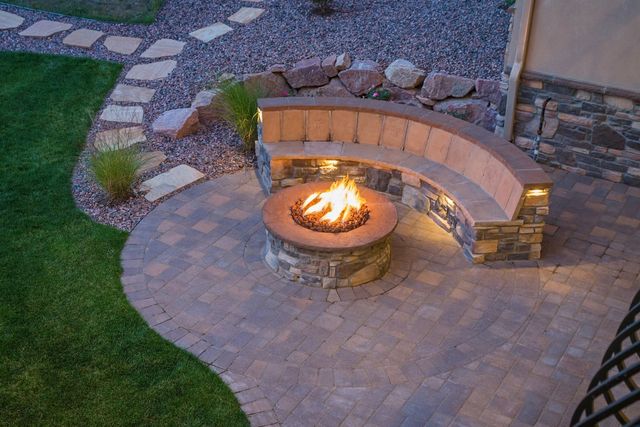 Backyard fire pit with curved stone bench seating, lit fire, and paved patio at dusk