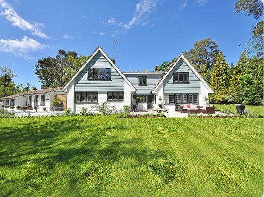 White farmhouse with dormer windows and red chairs, set behind a wide green lawn under a blue sky.