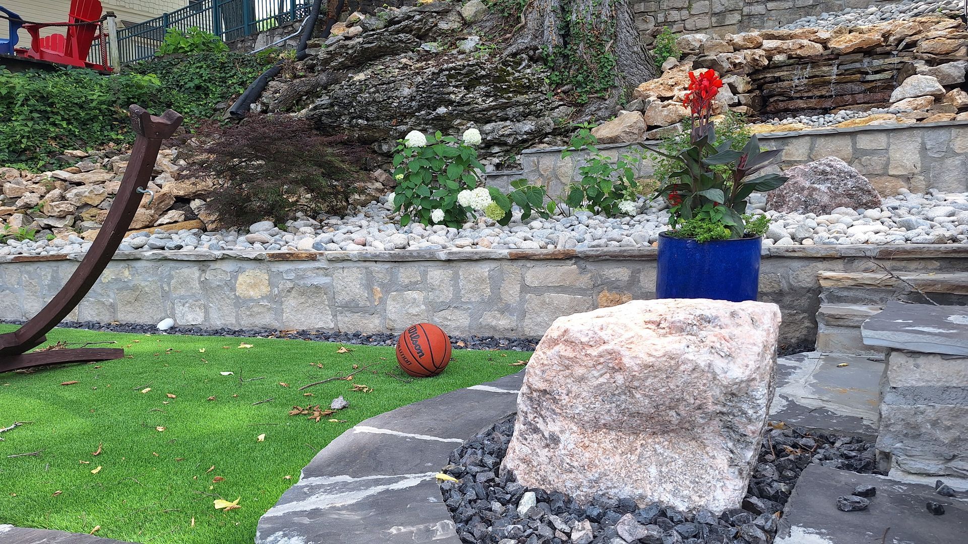 Garden with stone steps, a large rock in the foreground, and a blue pot with flowers beside a lawn.