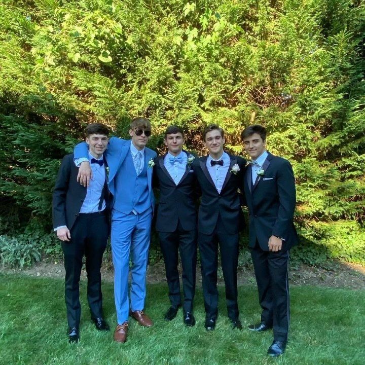 Five young men in formal attire pose outdoors. One wears a blue suit, the others wear black tuxedos; they smile at the camera.