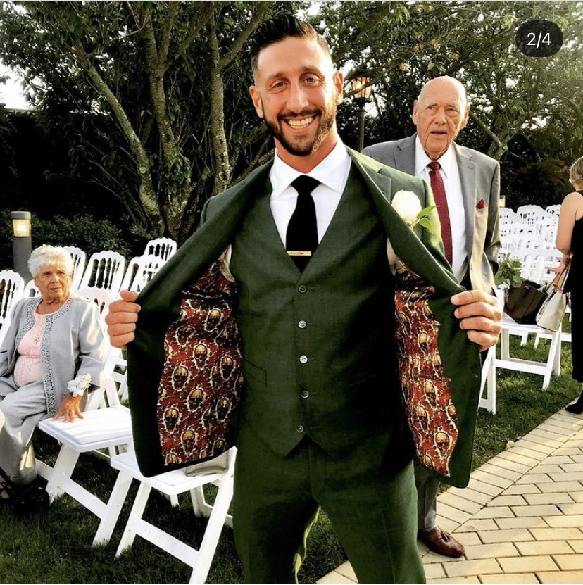 A man in a green suit with patterned lining, smiles while showing the inside of his jacket. An older man and woman are in the background.