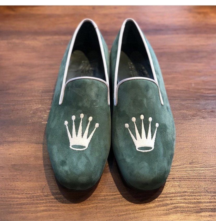 Pair of green suede loafers with a Rolex crown emblem embroidered on the toe of each shoe. White trim.