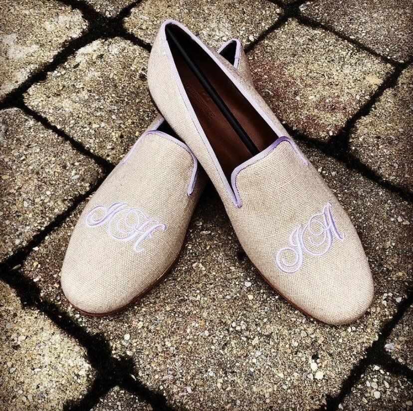 Pair of beige loafers with light purple monogram on a stone-tiled surface.