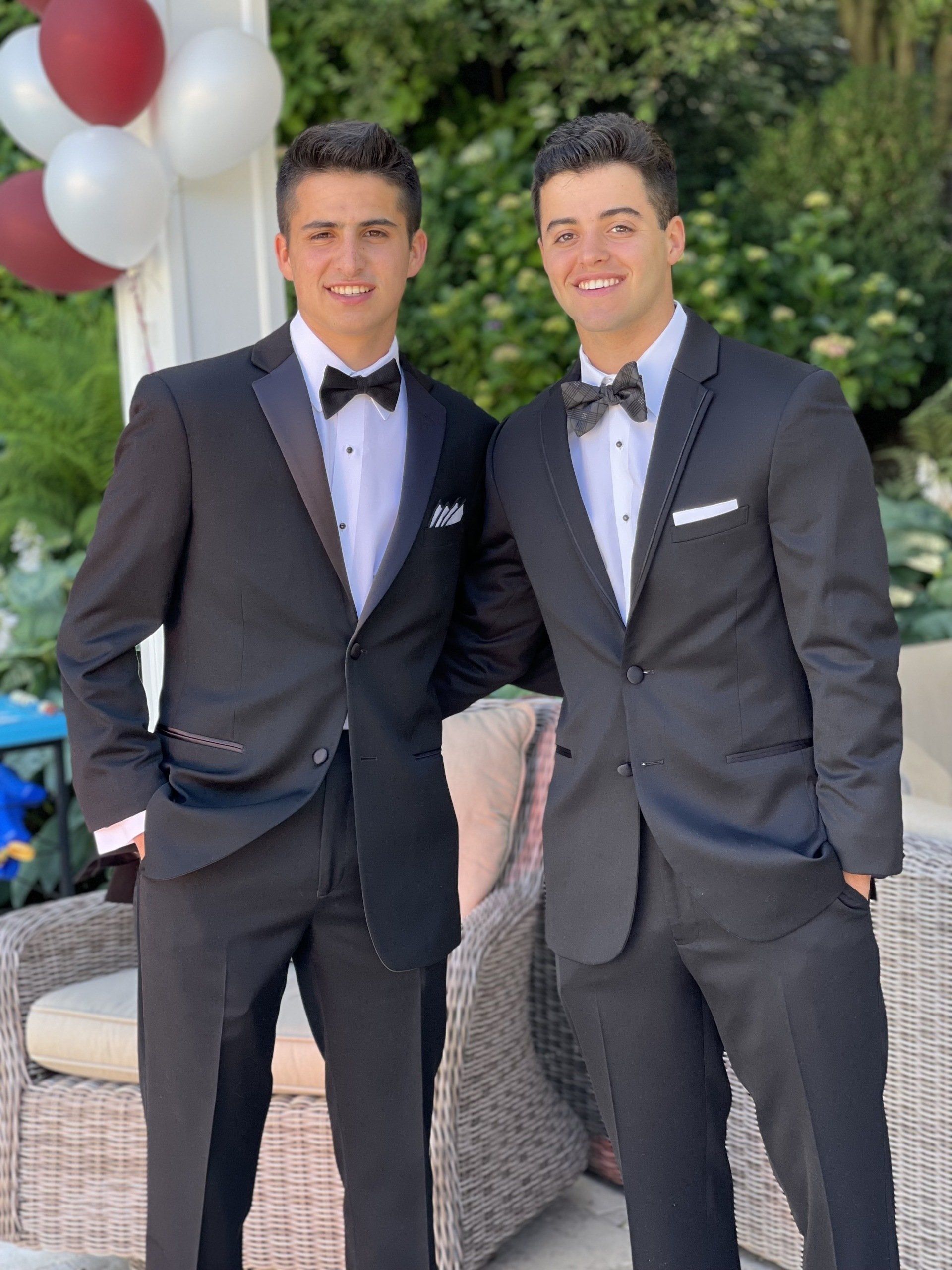 Two young men in black tuxedos pose outdoors, smiling at the camera. Balloons and greenery are in the background.