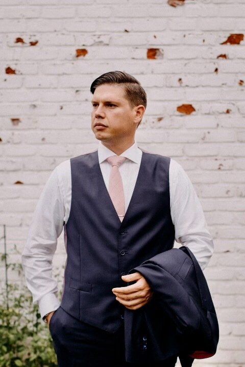 Man in navy vest, white shirt, and pink tie, holding a navy jacket, posing in front of a white brick wall.
