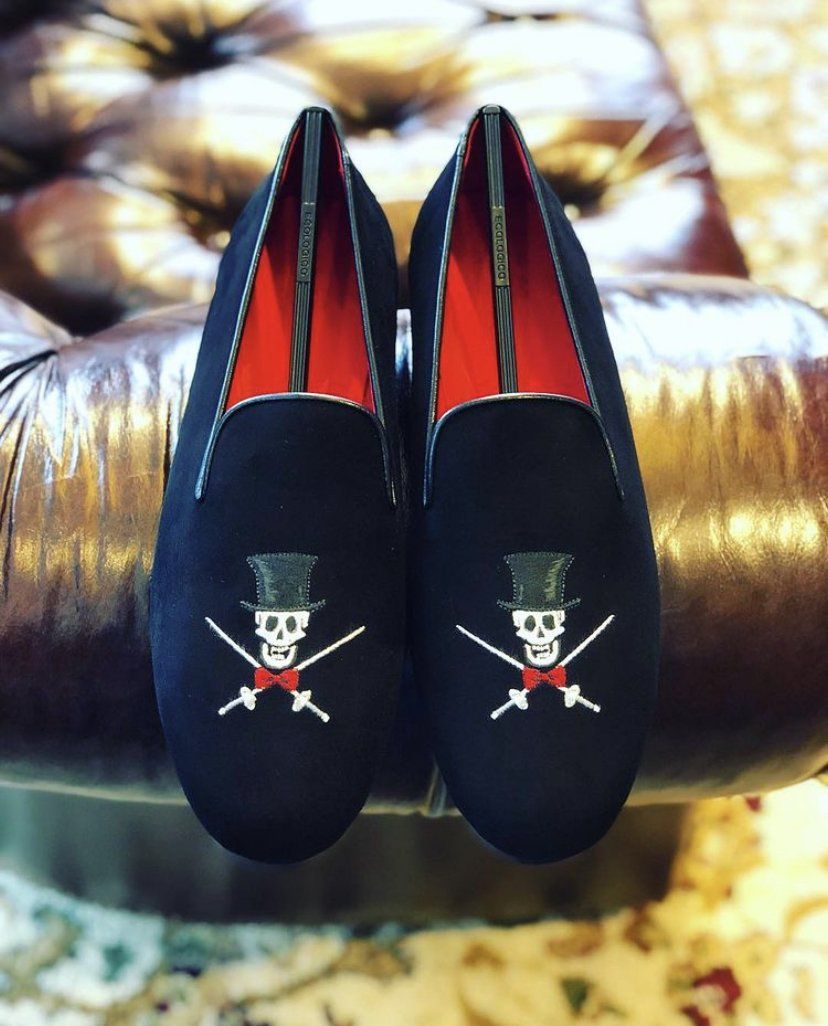 Black velvet loafers with a skull and crossbones design, red lining, resting on a brown surface.