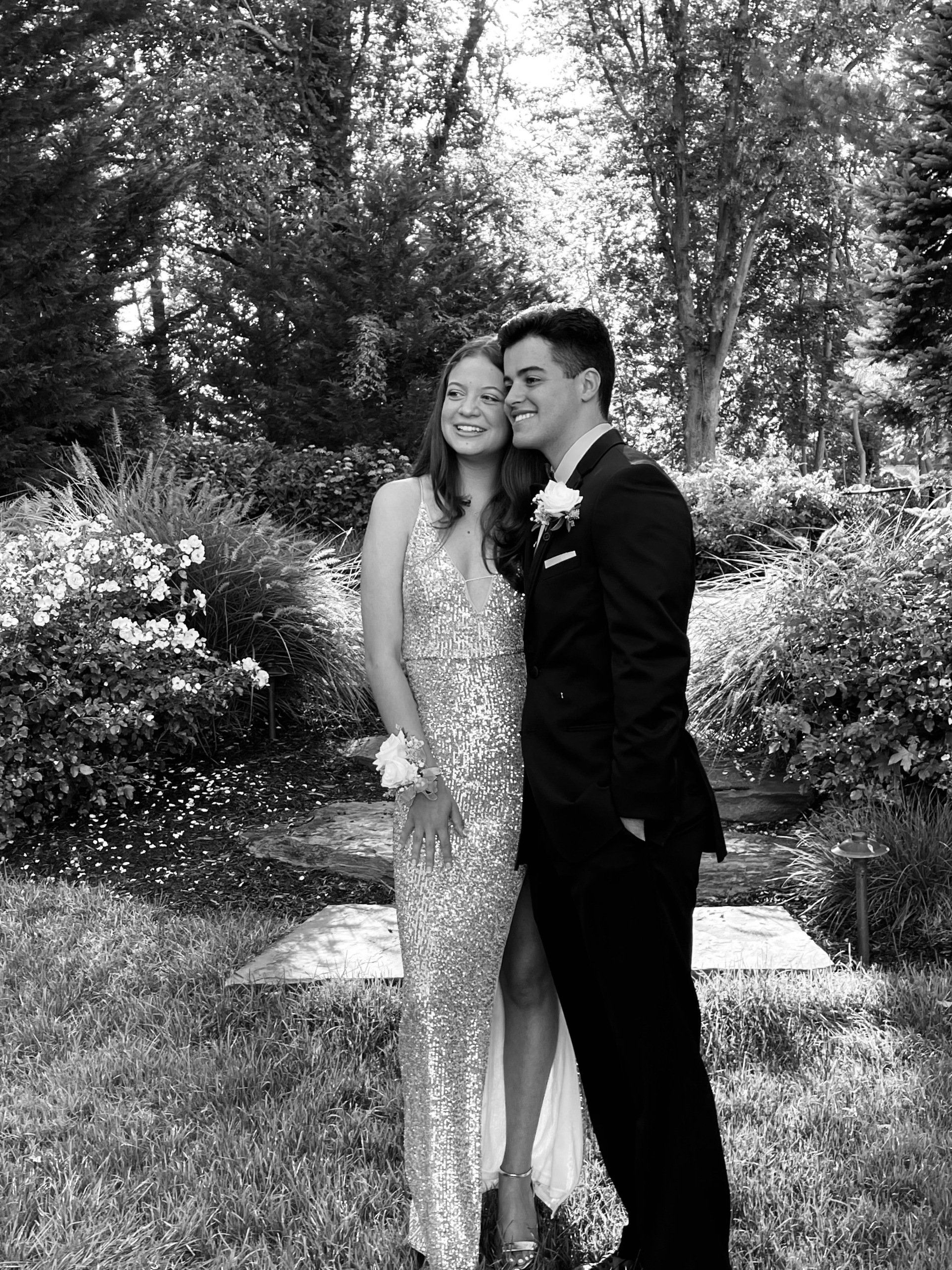 A couple dressed for prom poses in a garden. The woman wears a sequined gown, and the man is in a suit, smiling.