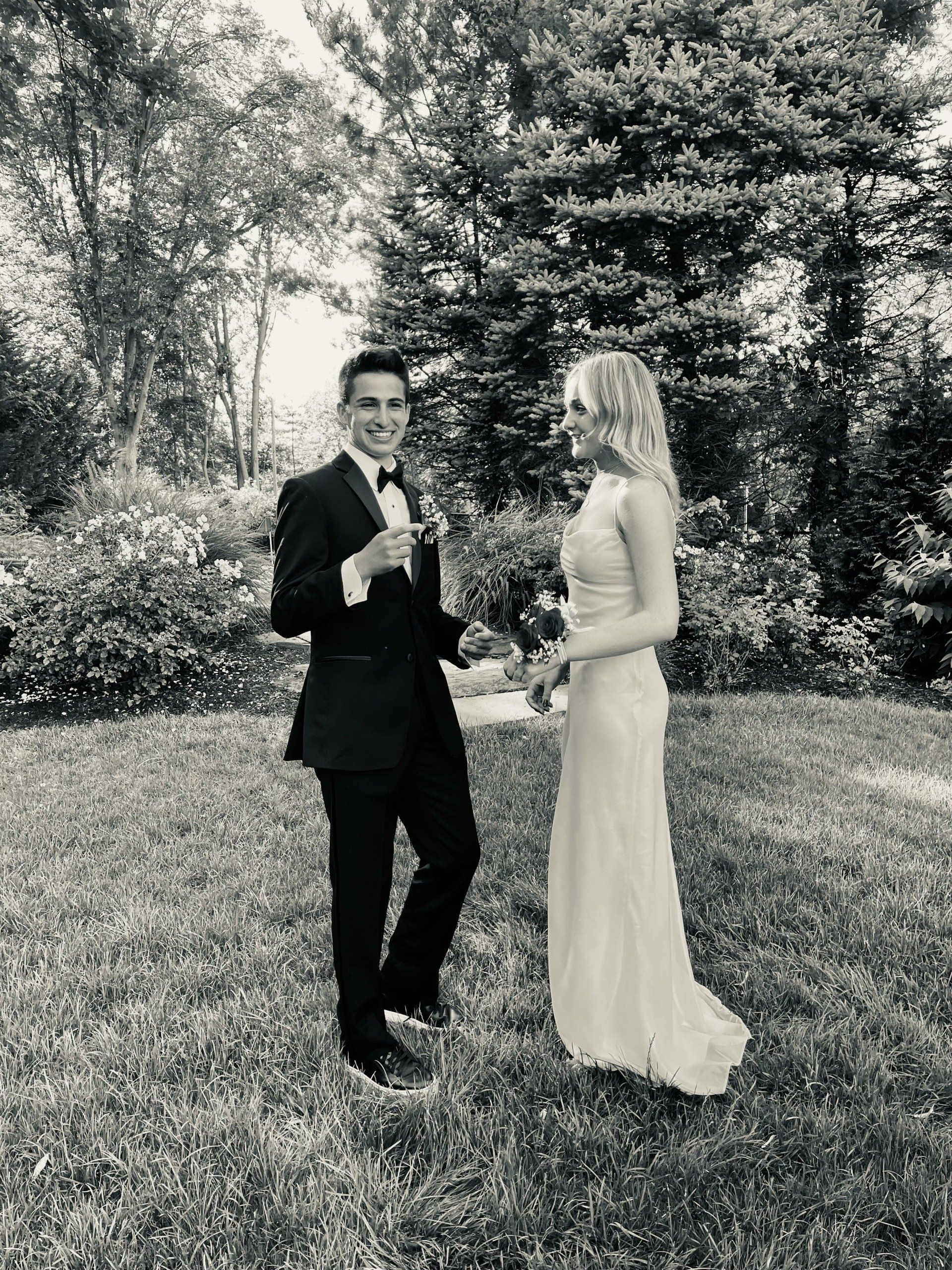 A well-dressed couple, a man and a woman, smile at the camera while standing on a grassy lawn with trees in the background.