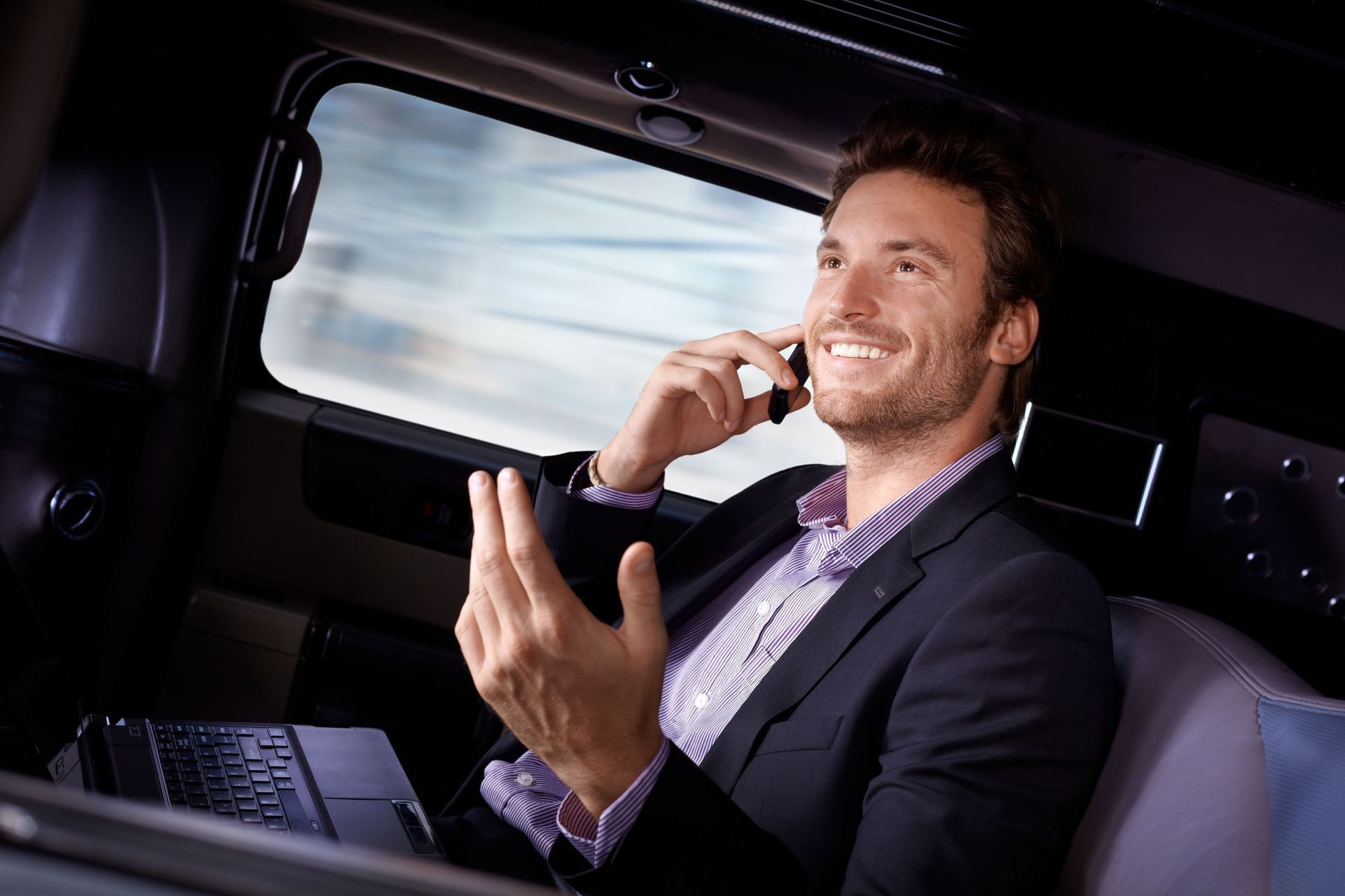 Man in suit talking on phone, smiling, in a vehicle. Laptop visible.