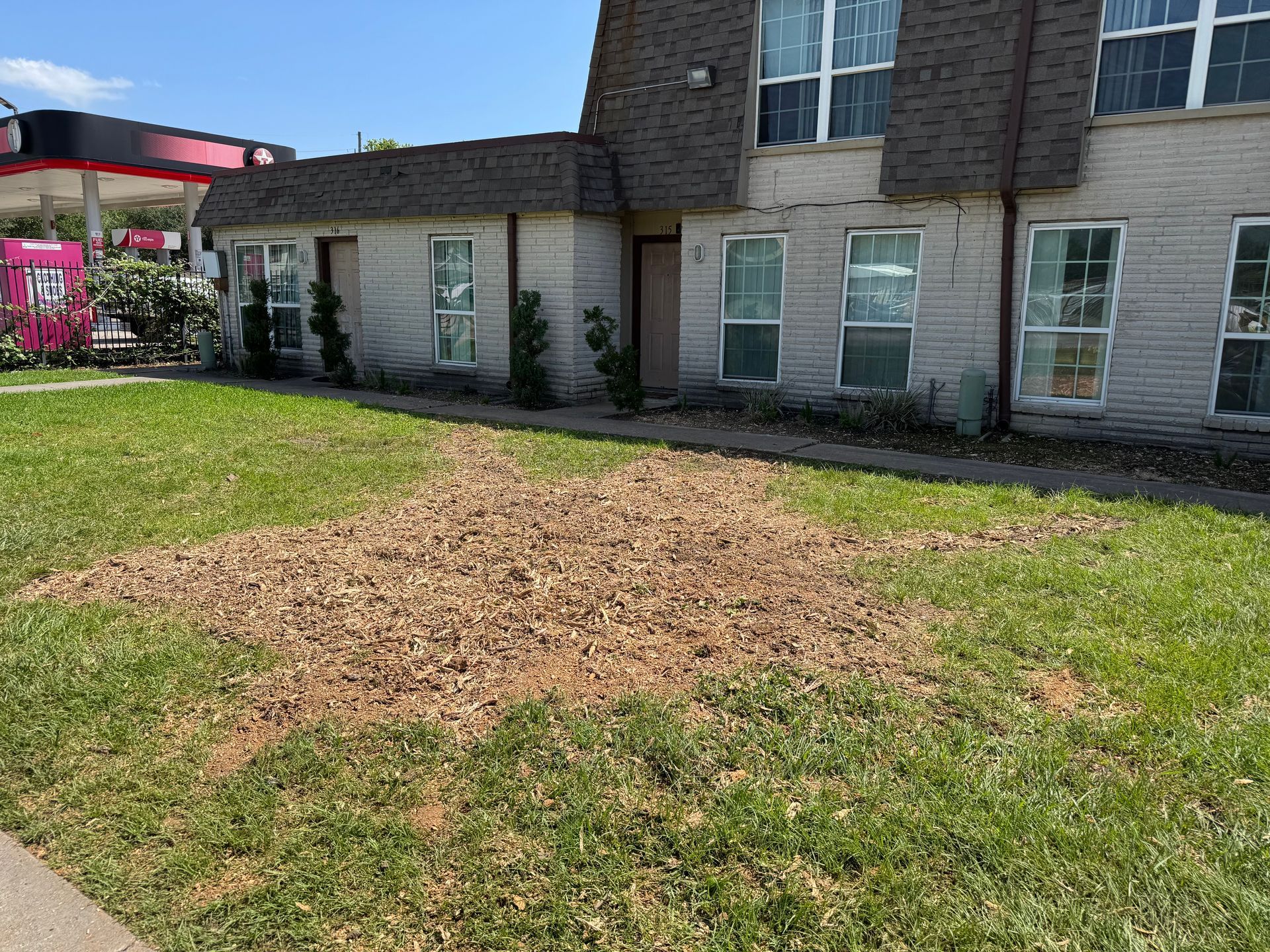 Brown patch of dirt in a grassy lawn in front of a two-story brick building with a gas station in the background.