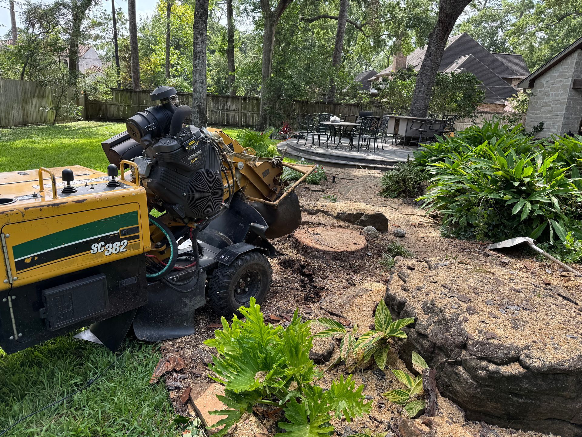 A yellow stump grinder in a backyard, grinding a tree stump near a patio.