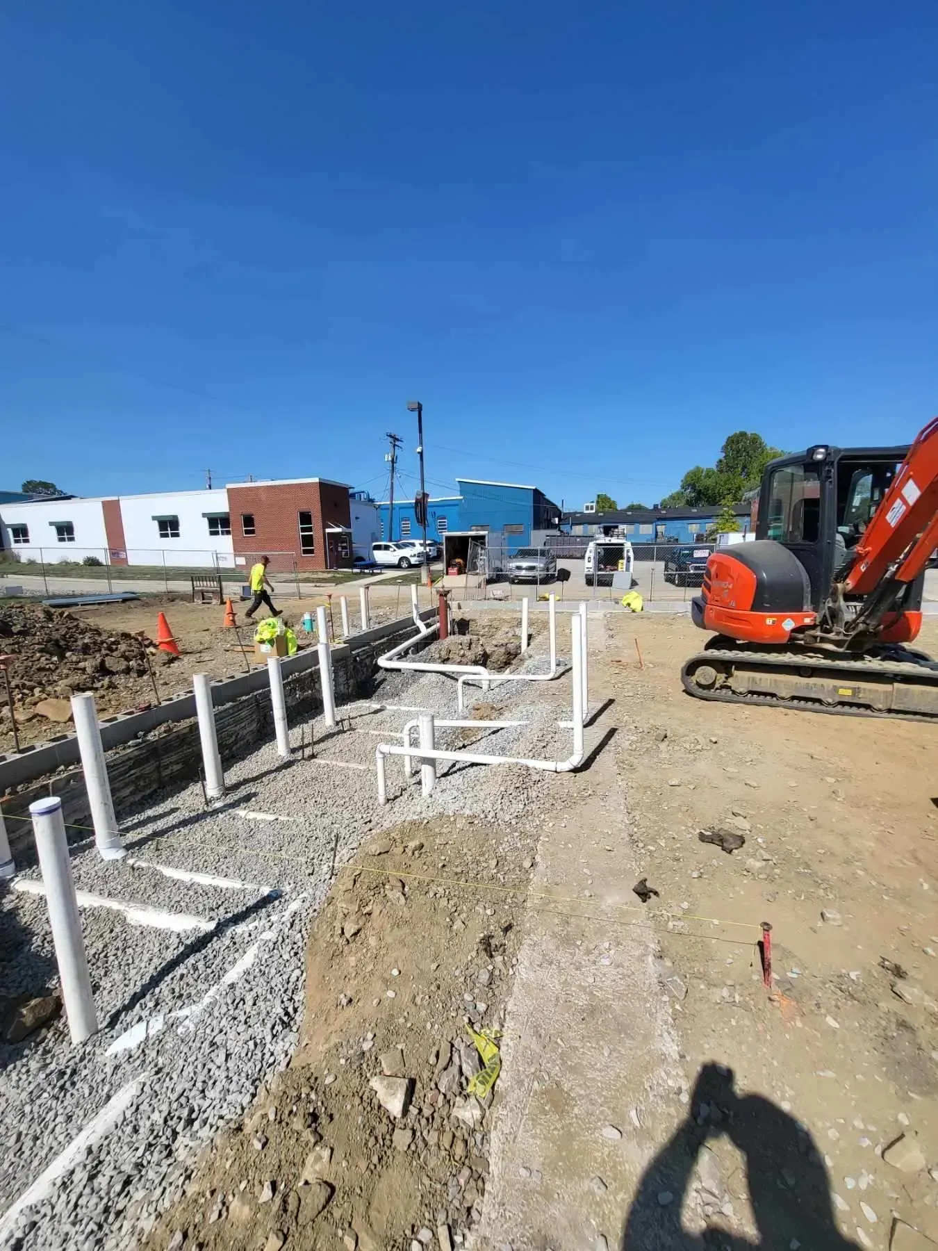 Construction site with piping, gravel, and a small excavator under a blue sky. Workers in vests.