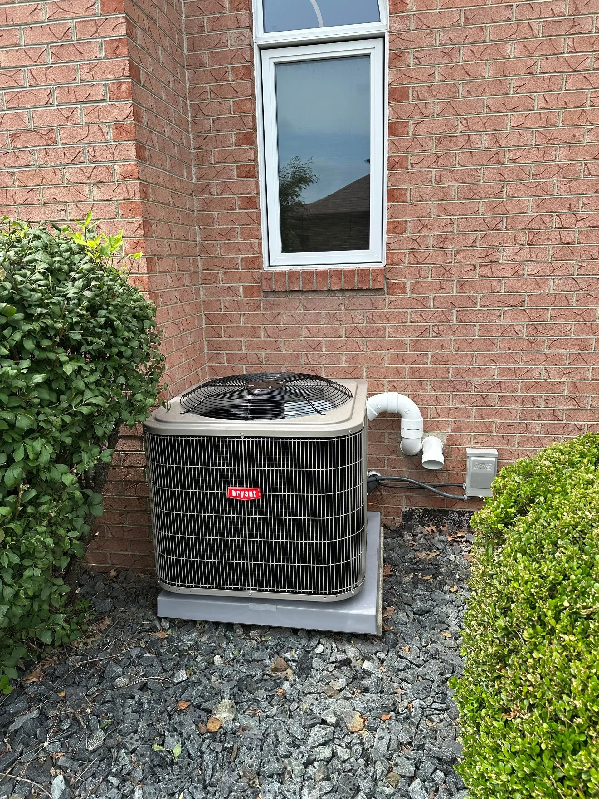 Air conditioning unit next to a brick wall, under a window, with bushes and rocks.
