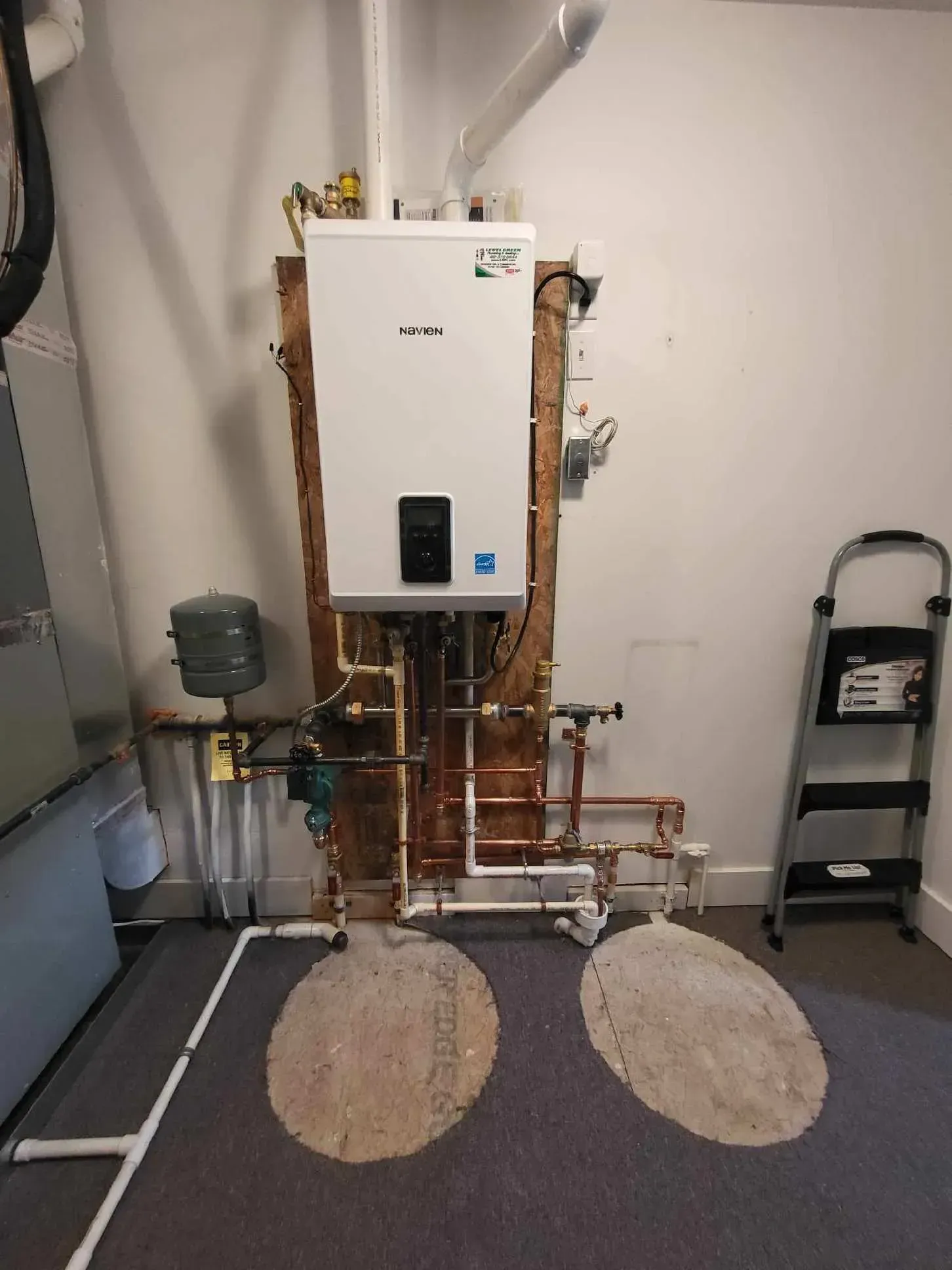 Water heater unit with copper pipes and gray step ladder in a room.