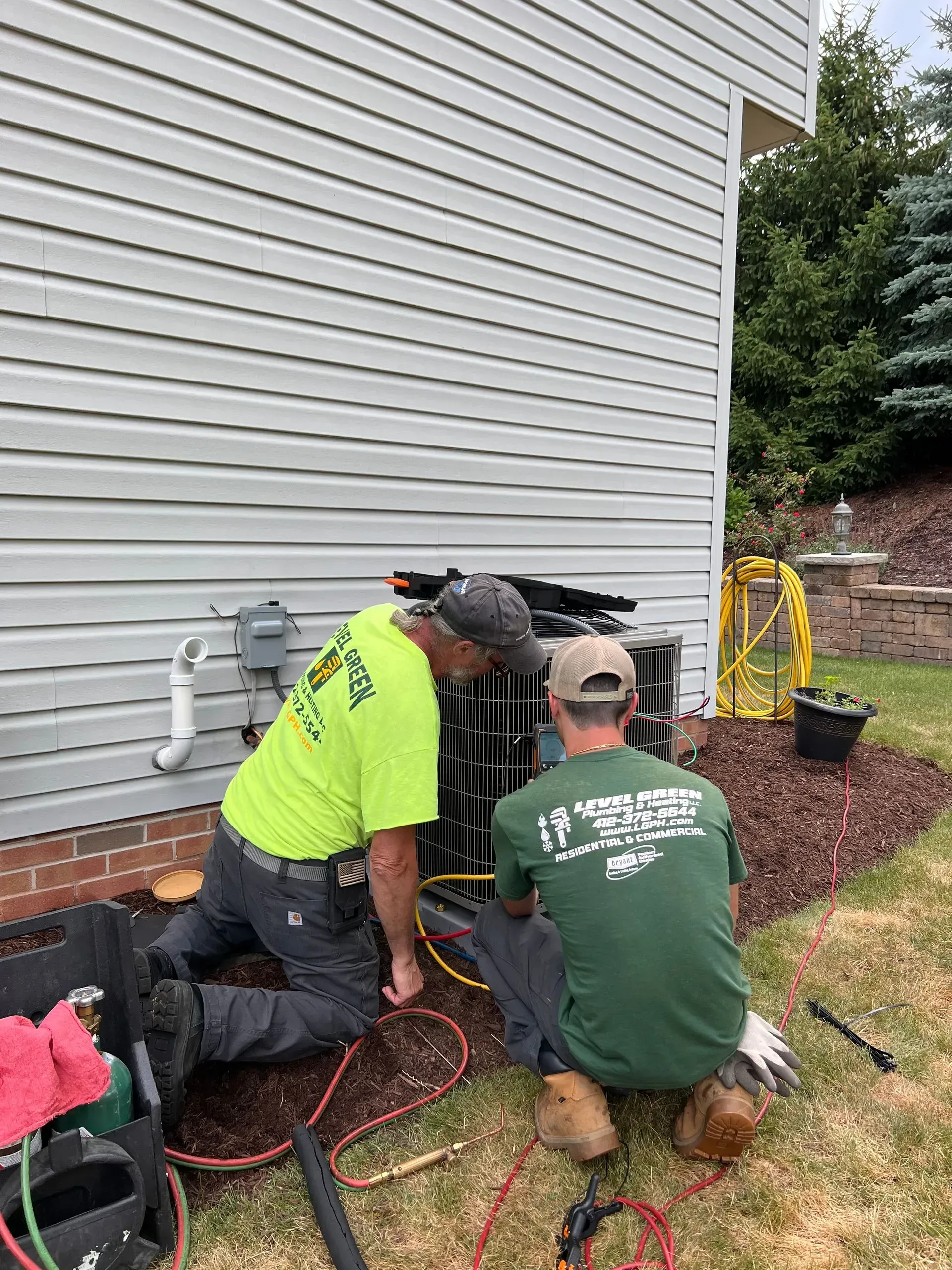 Two HVAC technicians working on an outdoor AC unit near a house. One kneels, the other sits; both inspect the unit.