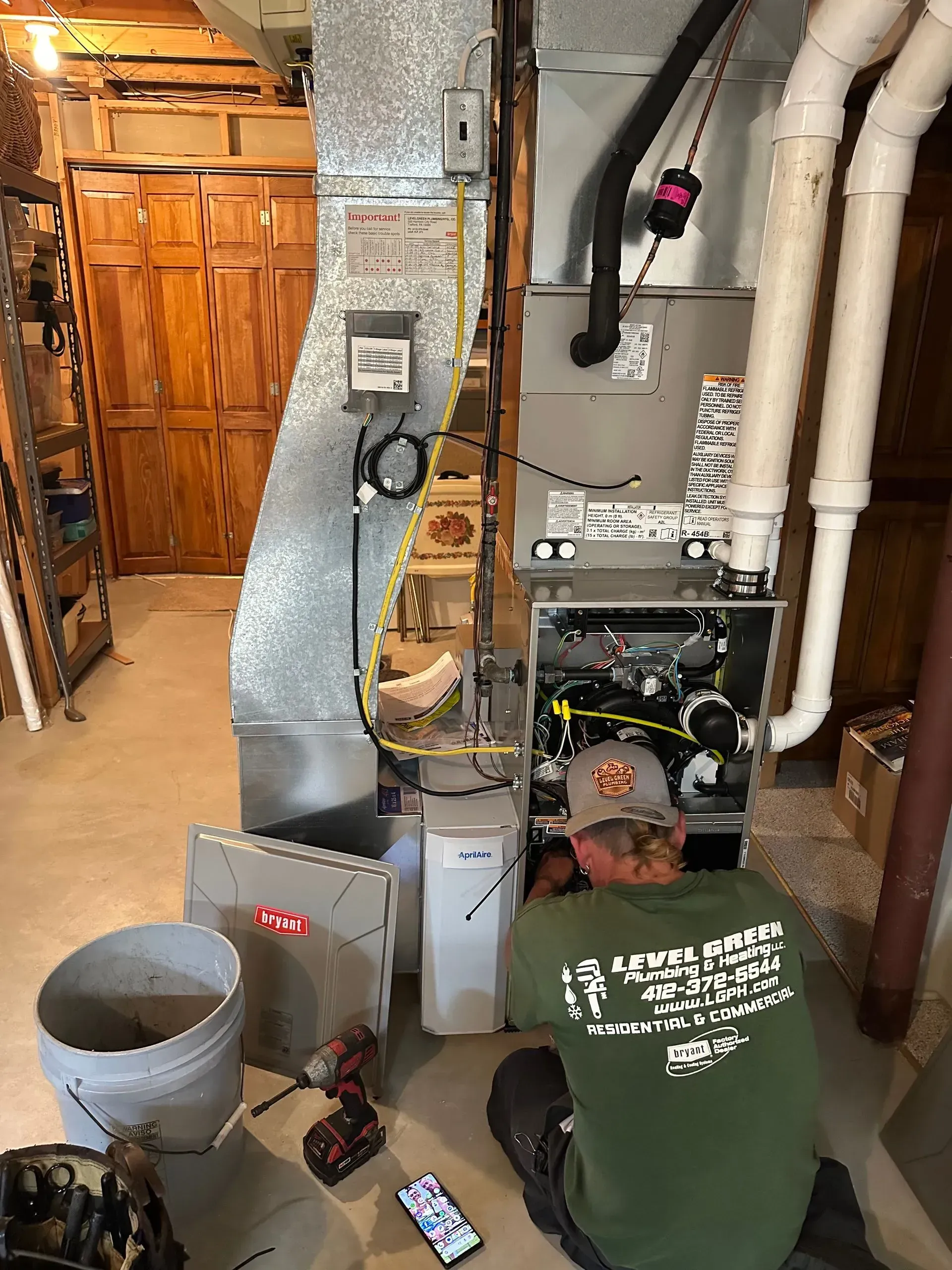HVAC technician working on a furnace in a basement. Man in green shirt, tools, gray and white components.