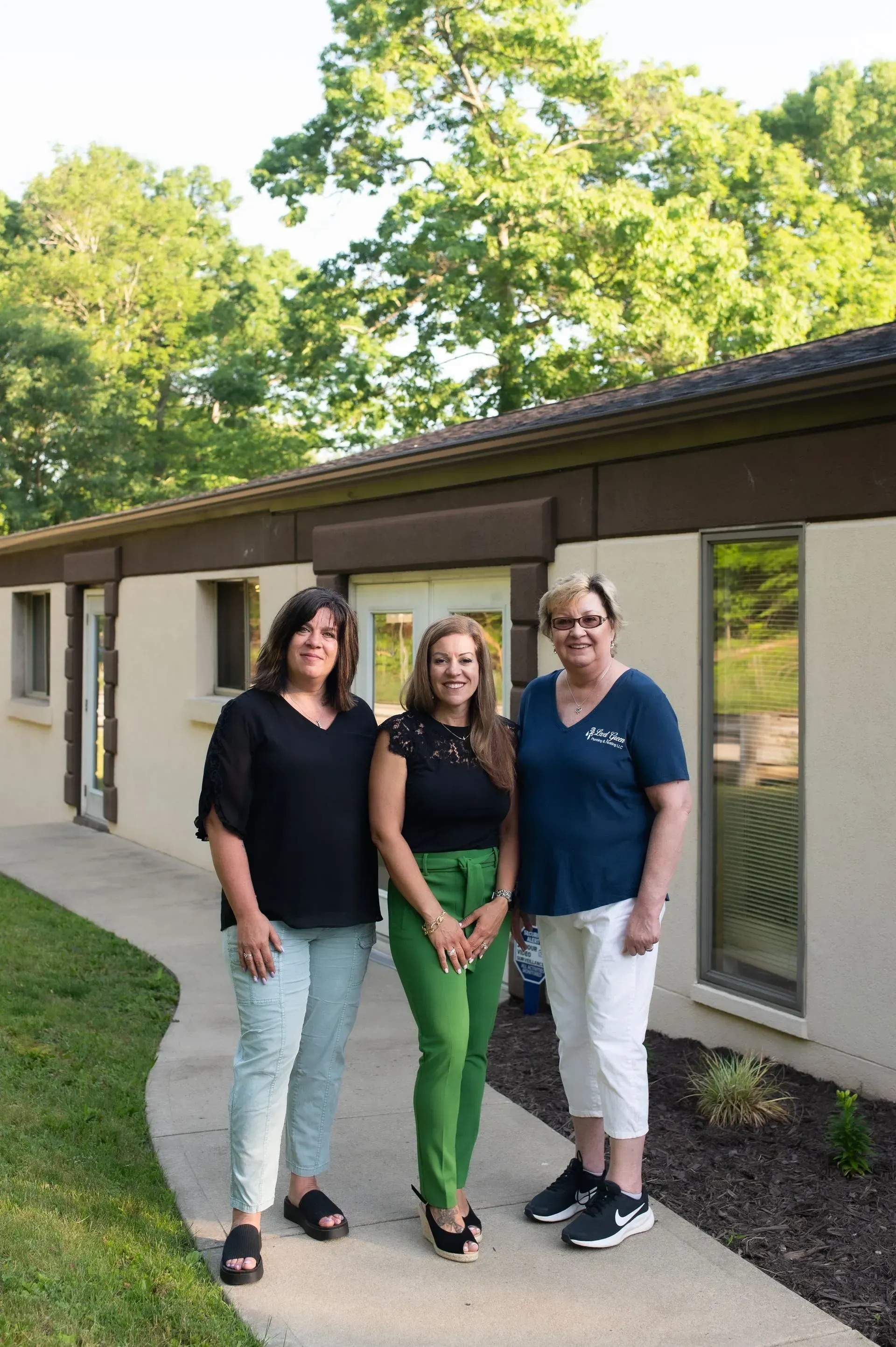 Three women stand outside a building; one in green pants, others in black tops and patterned/white pants.