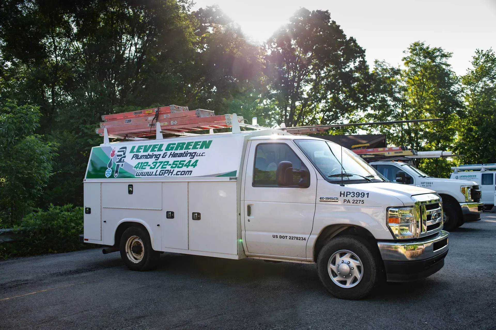 White work van with company logo parked outdoors; sunlight filters through trees in background.