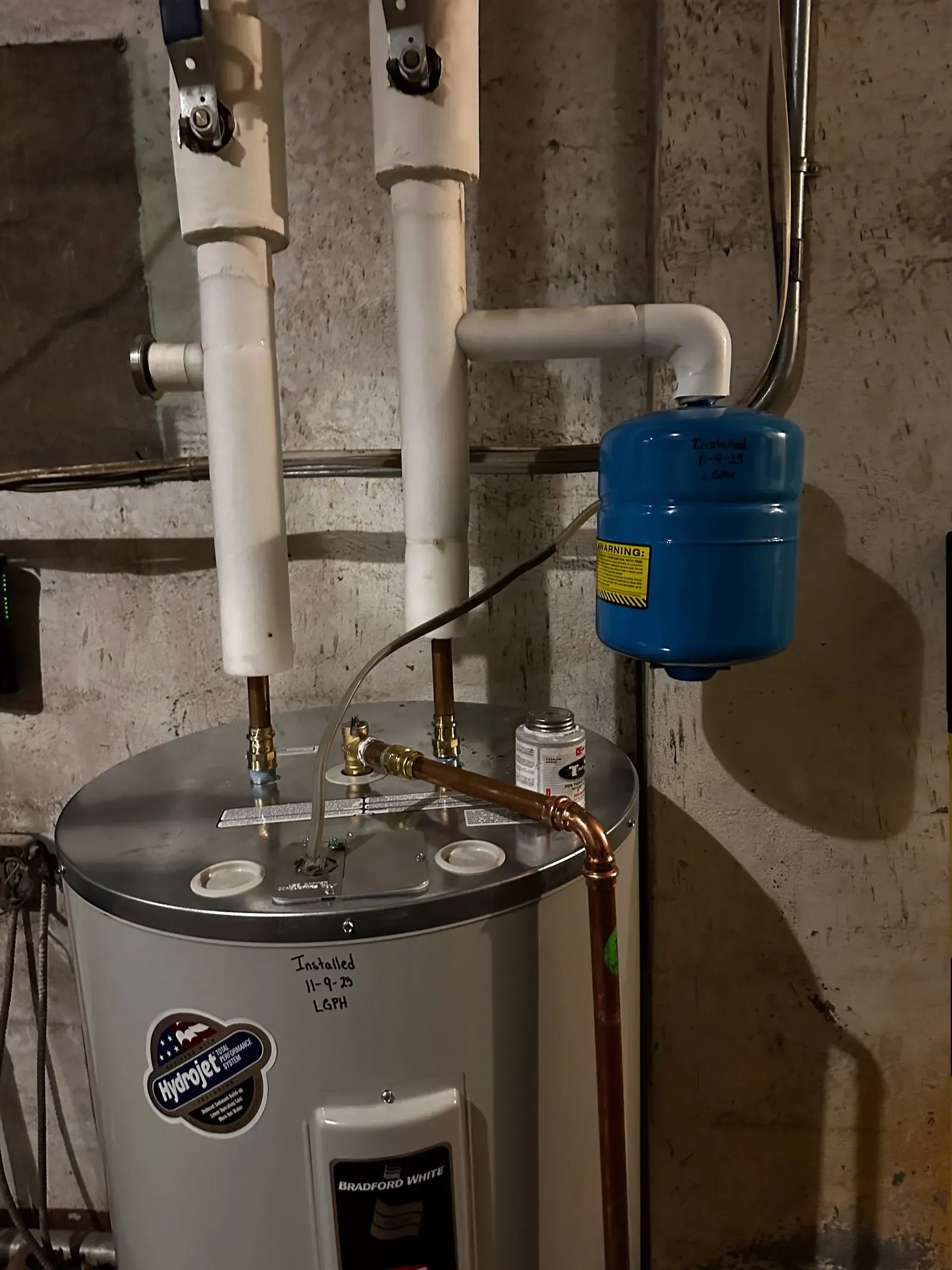 A water heater with white pipes, copper tubes, and a blue expansion tank in a basement.