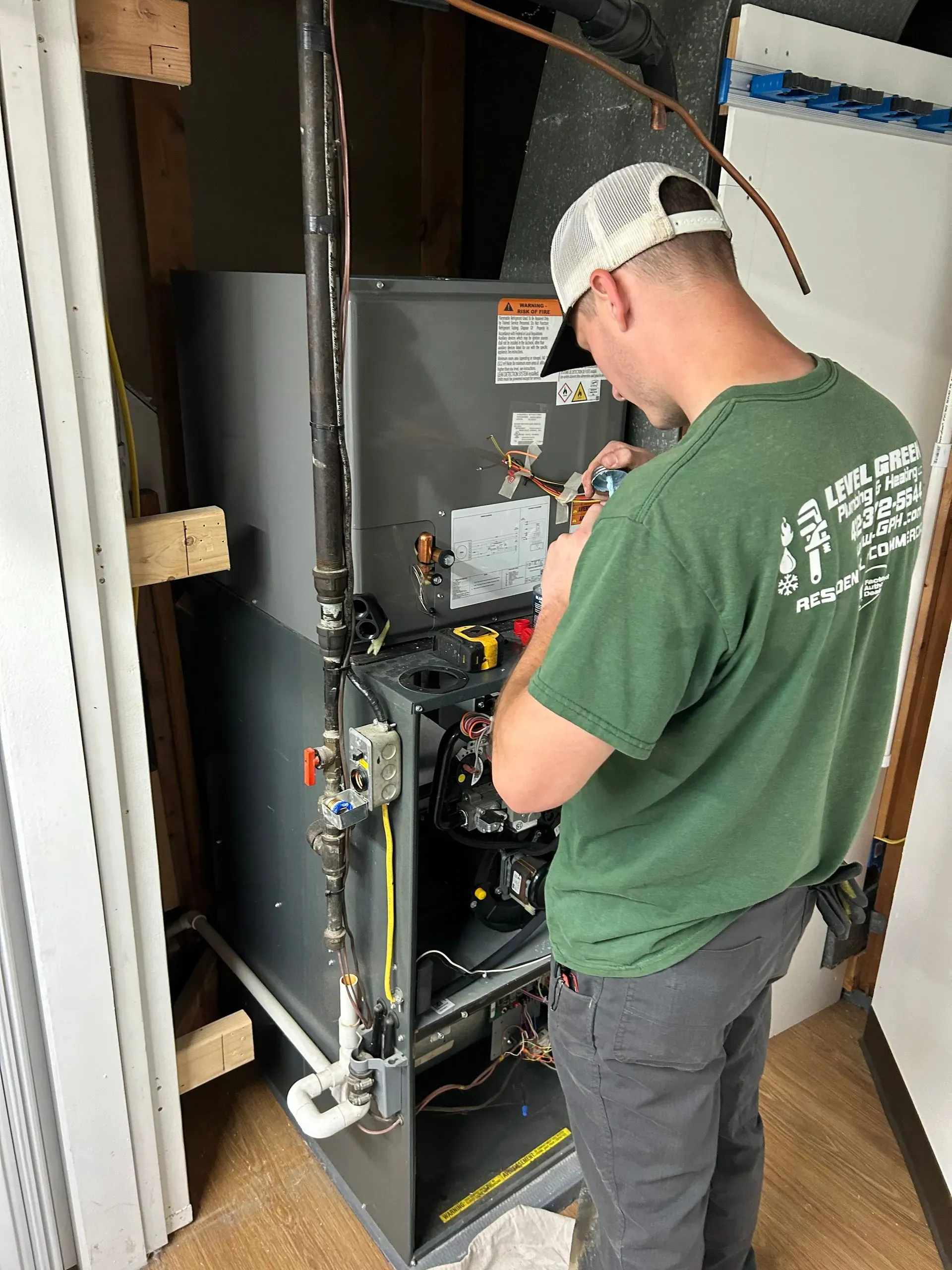 Man in green shirt repairs HVAC unit. Indoors, he is working on the internal components with a neutral expression.