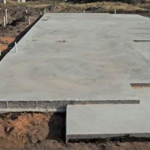 Concrete foundation slab, rectangular shape, sits on dirt. Pipes protrude from slab edges, suggesting construction site.