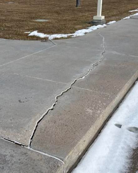 Cracked concrete sidewalk with edge, snow patches, and brown grass in the background.