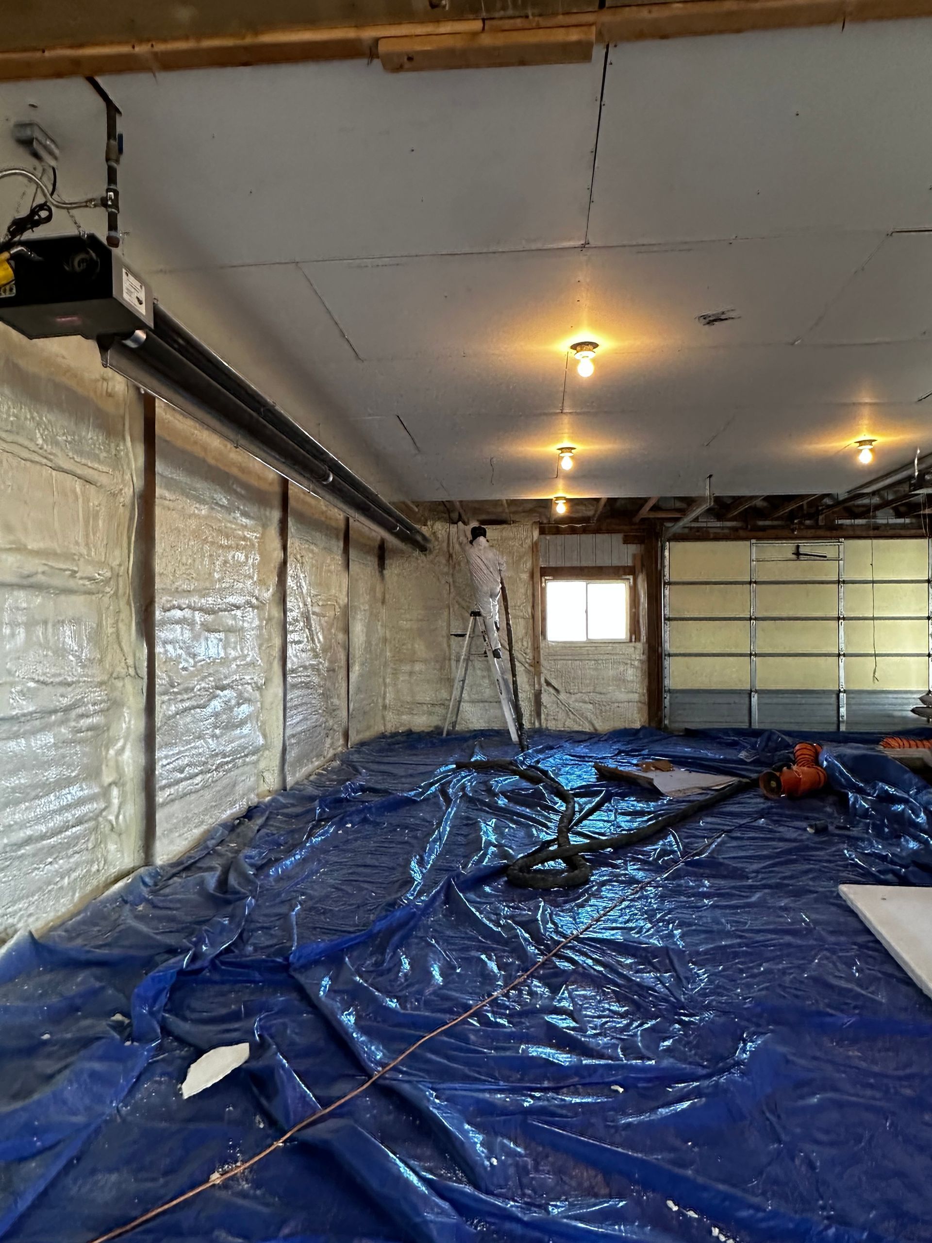 Garage interior with spray foam insulation, blue tarp covering floor, and overhead lights.