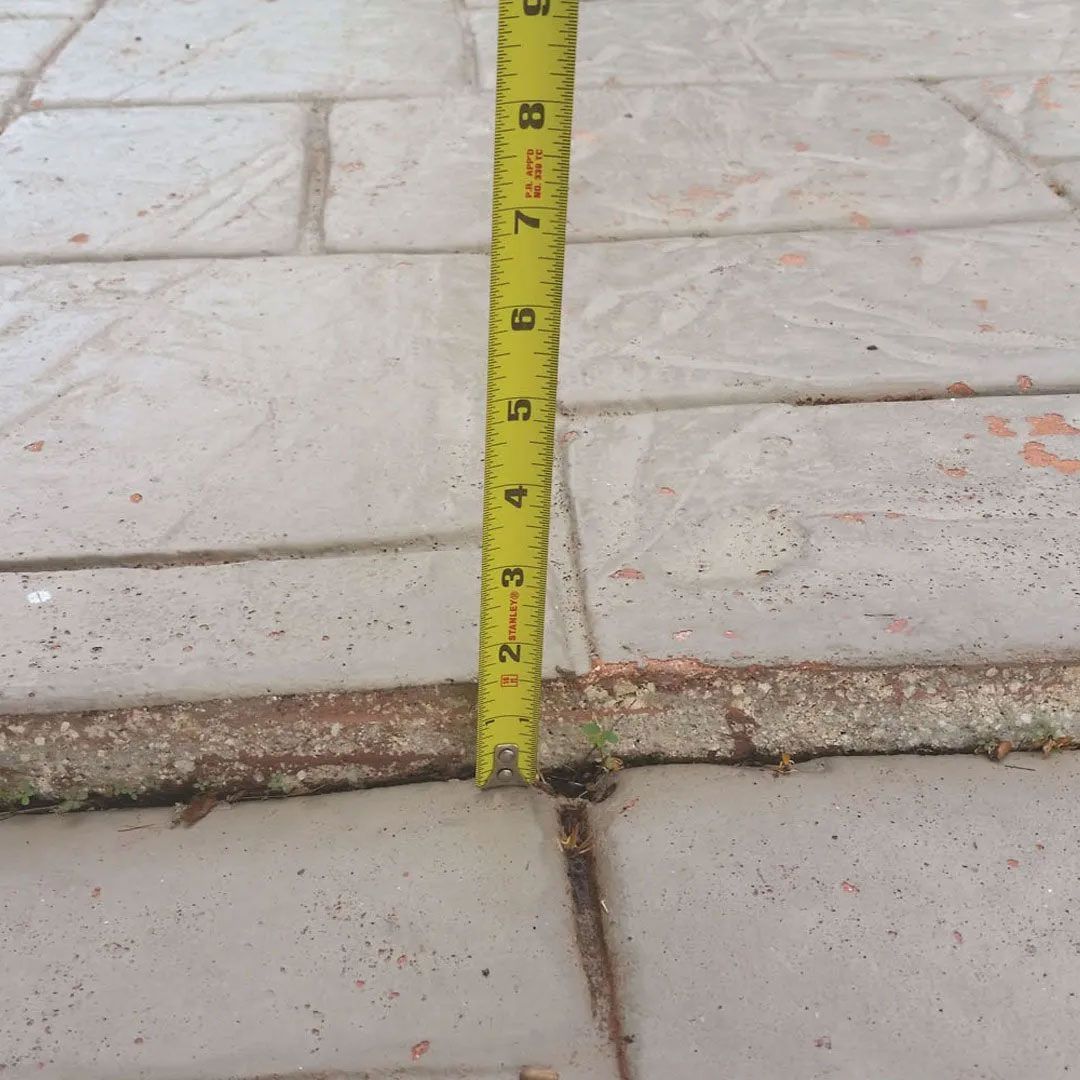 A yellow tape measure is placed vertically over a crack in a concrete patio, showing about 3 inches.