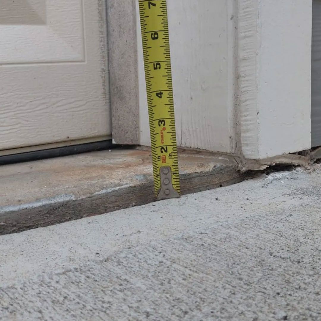 A measuring tape shows a 2-inch height difference between a concrete floor and a doorway threshold.