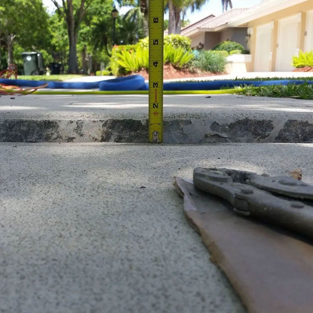 A tape measure shows a concrete slab's height difference. Pliers are in the foreground, and houses are in the background.