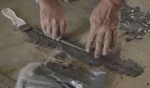 Hands using a notched trowel to spread gray mortar on a light-colored surface.