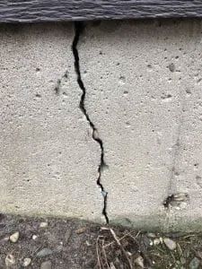 Vertical crack in a concrete foundation wall; dark gray roof above; ground with pebbles and dry grass below.