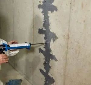Person sealing a vertical crack in a concrete wall with a caulking gun, gray sealant visible.
