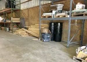 Warehouse interior with shelving, barrels, pallets, and lumber on a concrete floor.
