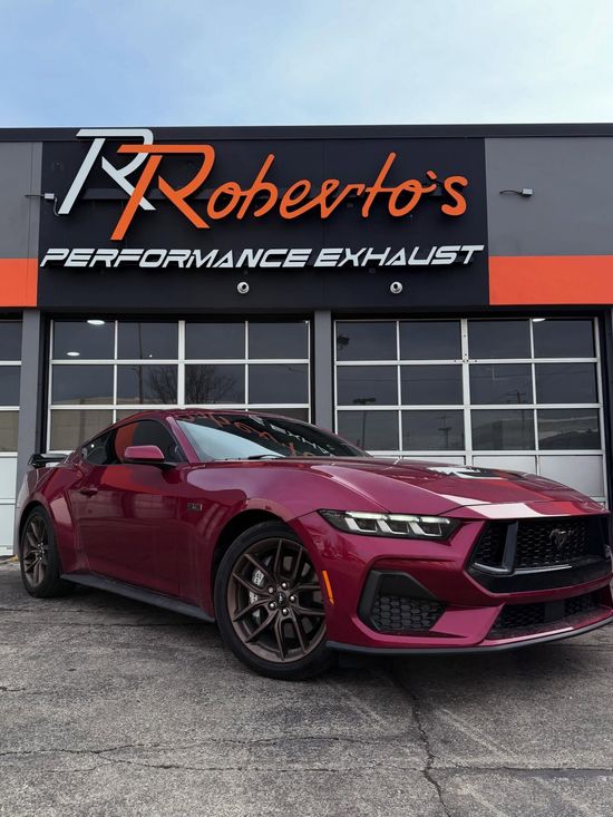 A deep red Ford Mustang parked in front of a building with a Roberto’s Performance Exhaust sign.