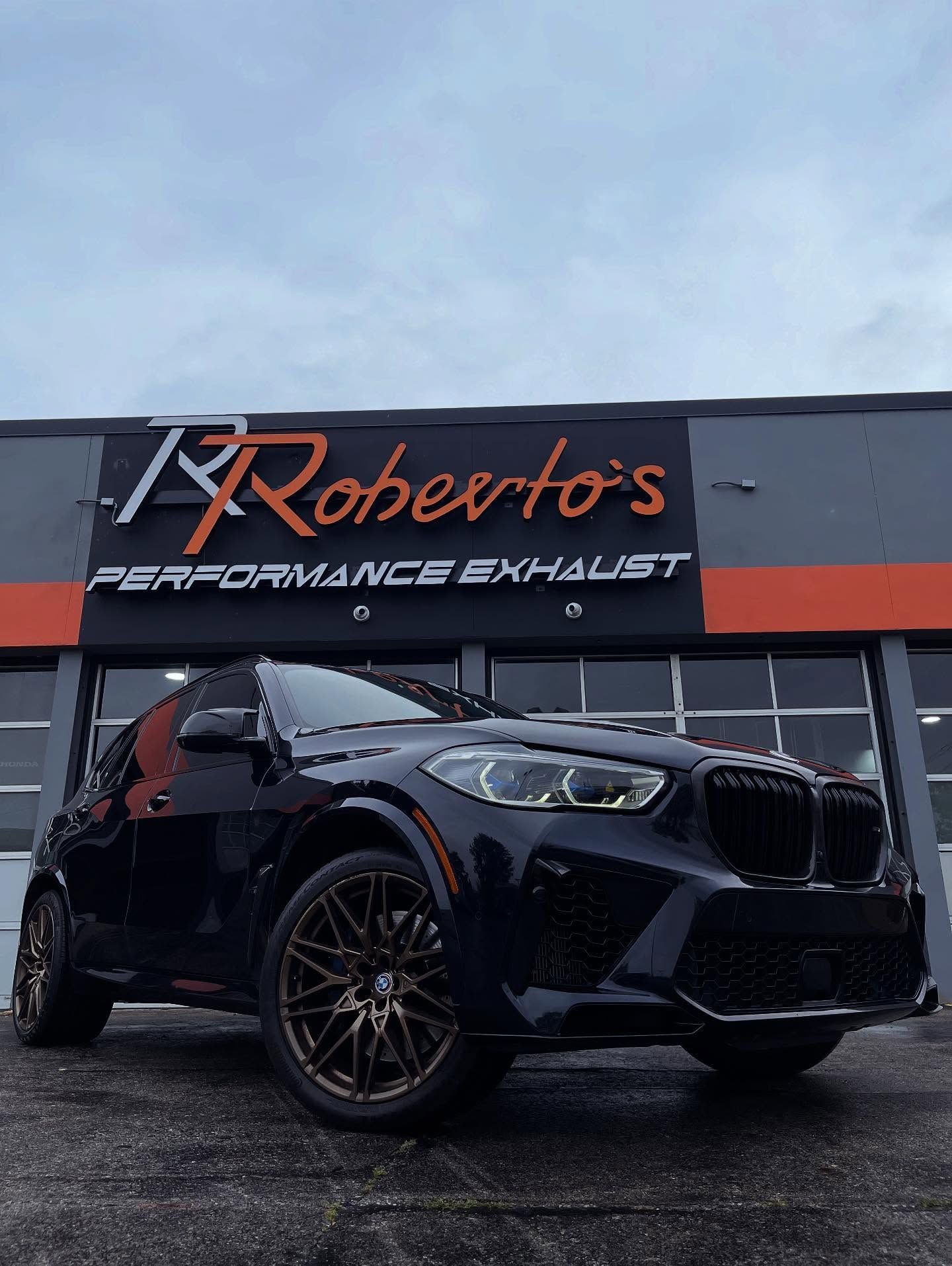 A dark BMW SUV with bronze wheels parked outside Roberto's Performance Exhaust shop under a cloudy sky.