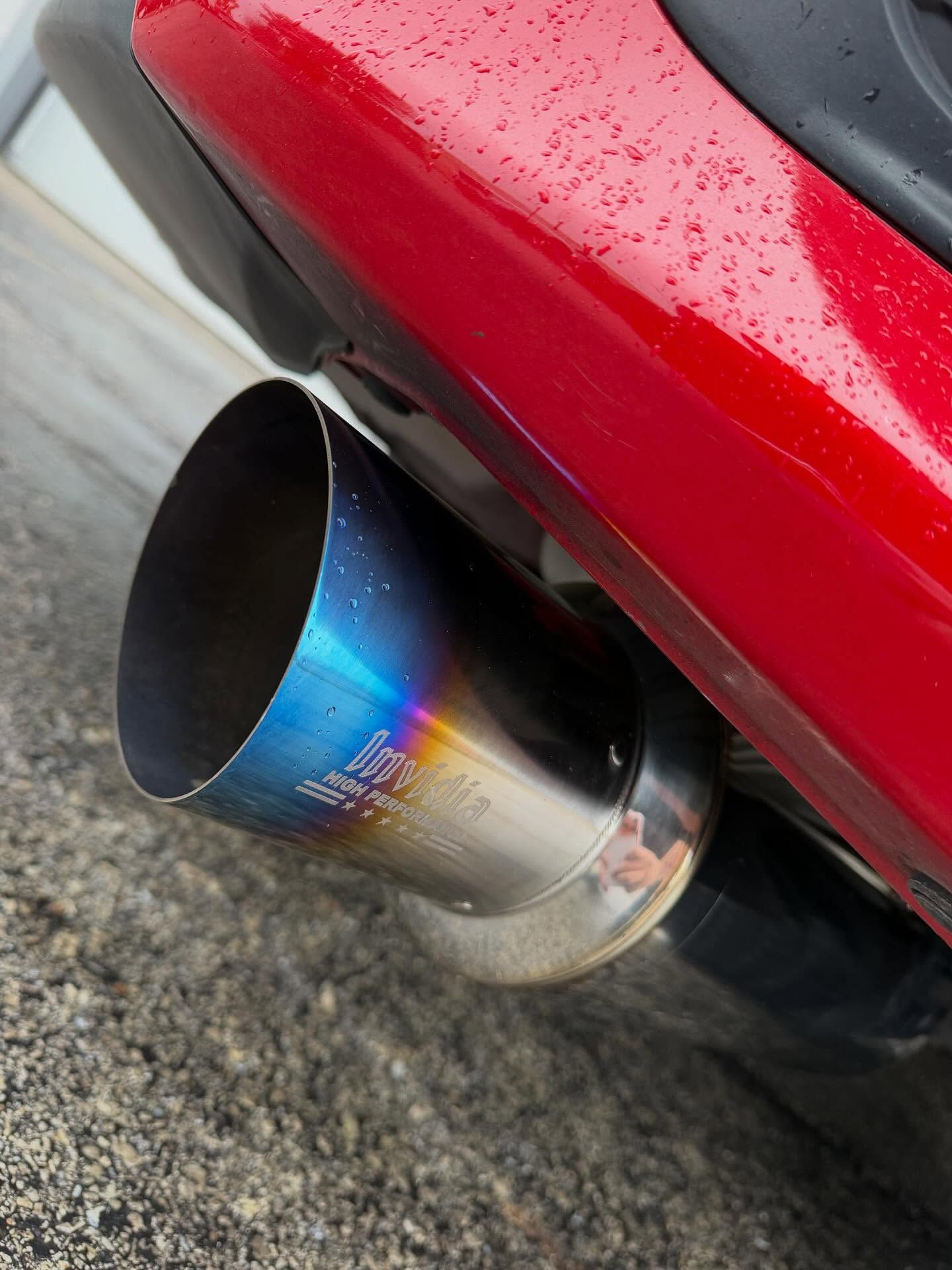 A close-up shot of a stainless steel car exhaust tip with a blue and gold burnt-finish, mounted on a red car bumper.