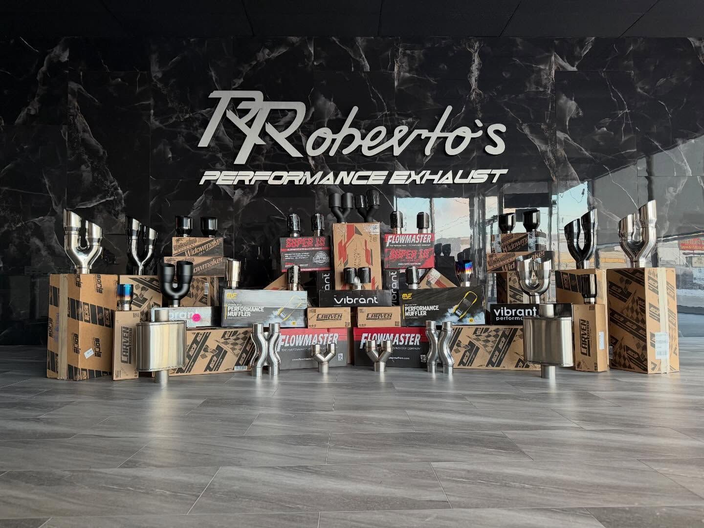 A display of various metal exhaust components and branded boxes arranged in front of a Roberto's Performance Exhaust sign.