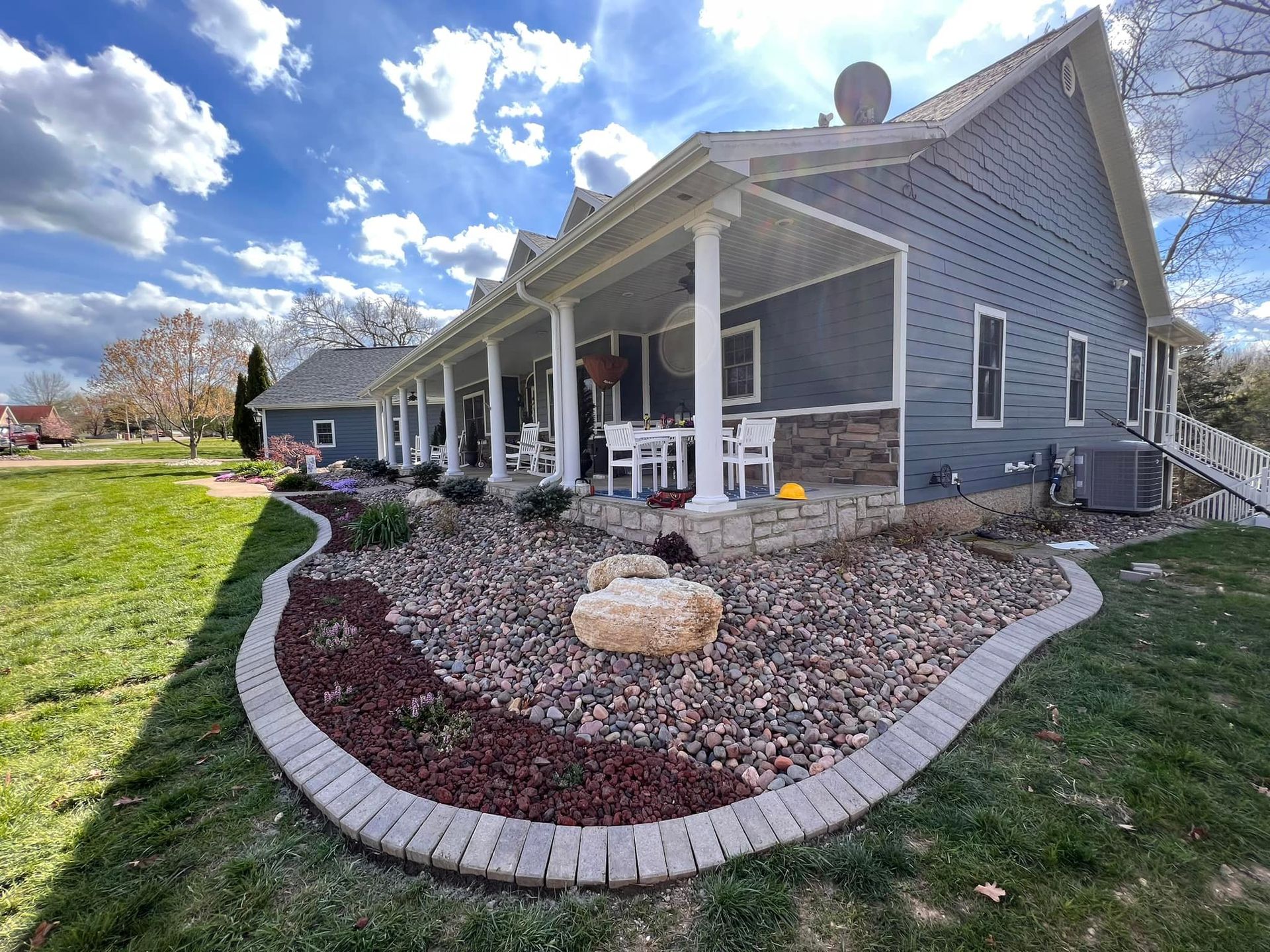 Blue house with a long porch, rock garden, and green lawn under a cloudy sky.