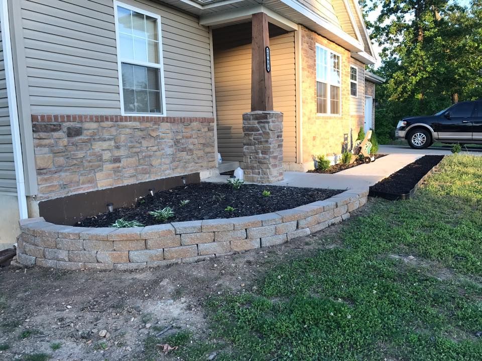 Curved retaining wall with flower bed near a house's entrance, with a dark-colored truck parked nearby.
