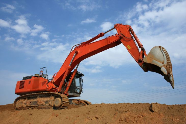Orange excavator on a dirt mound, its bucket raised against a blue sky.
