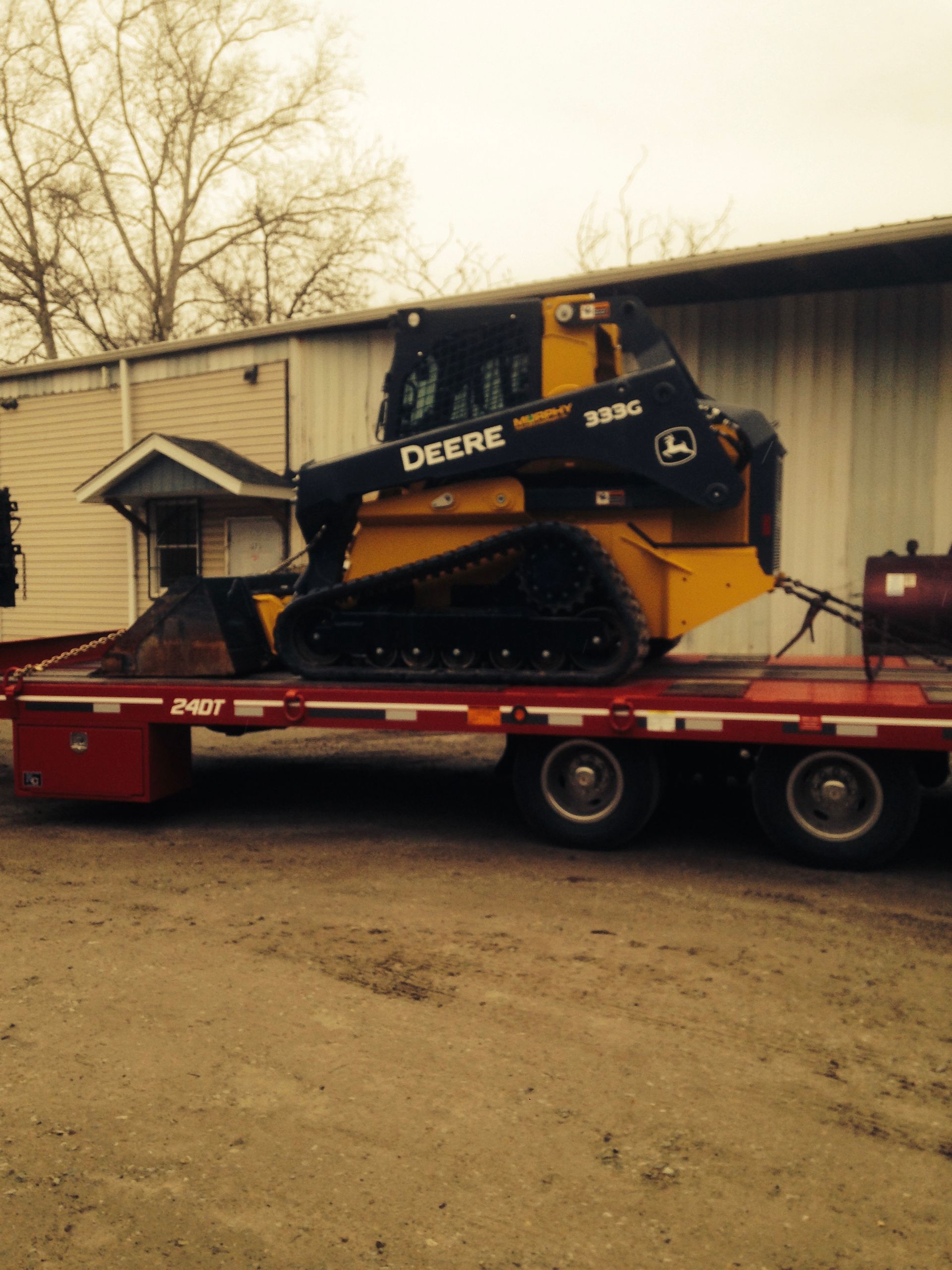 Yellow John Deere skid steer on a red trailer beside a building in a muddy yard
