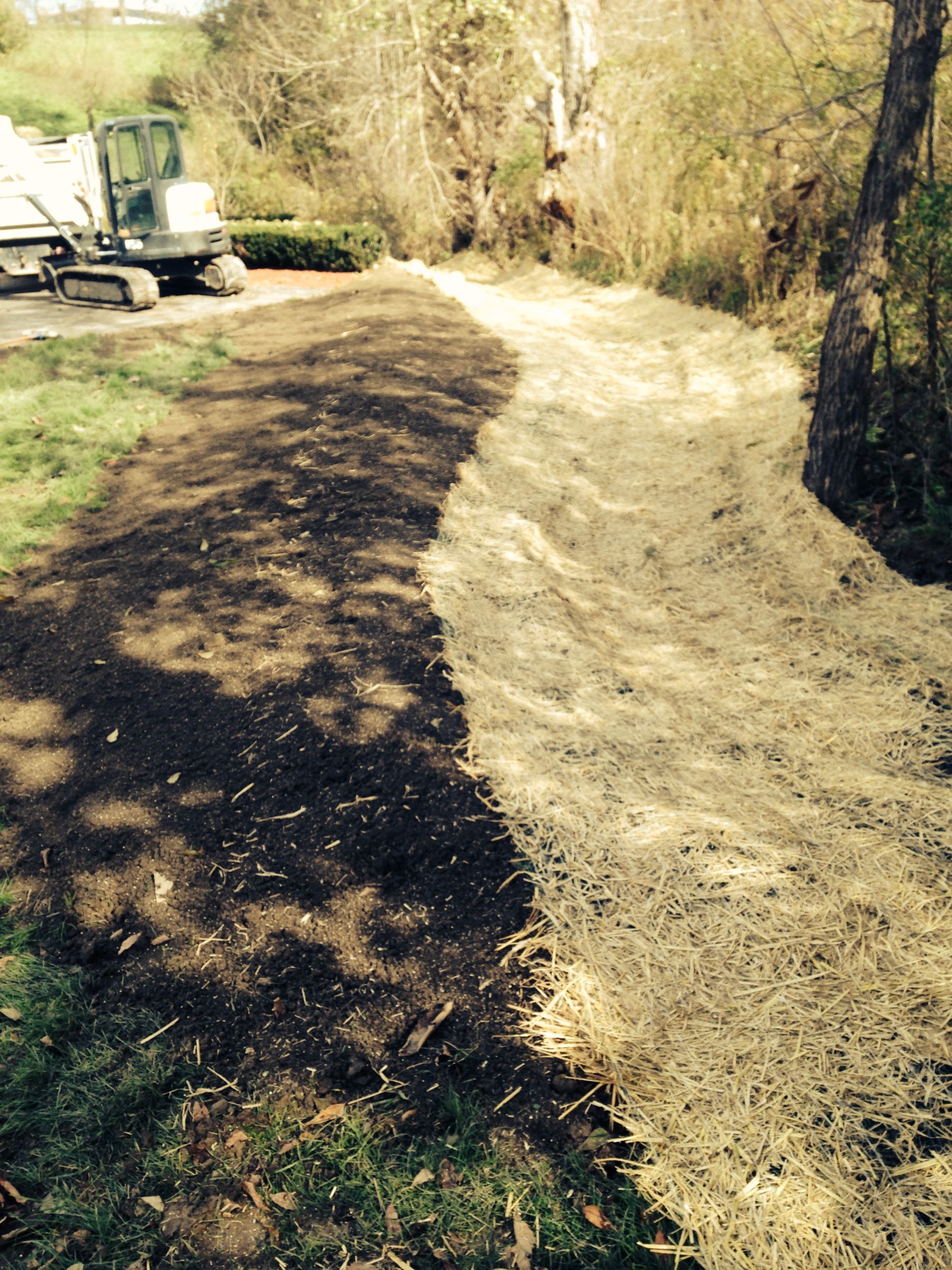 Dirt path beside a wooded creek, with a small excavator on the left and fresh pale gravel along the right side