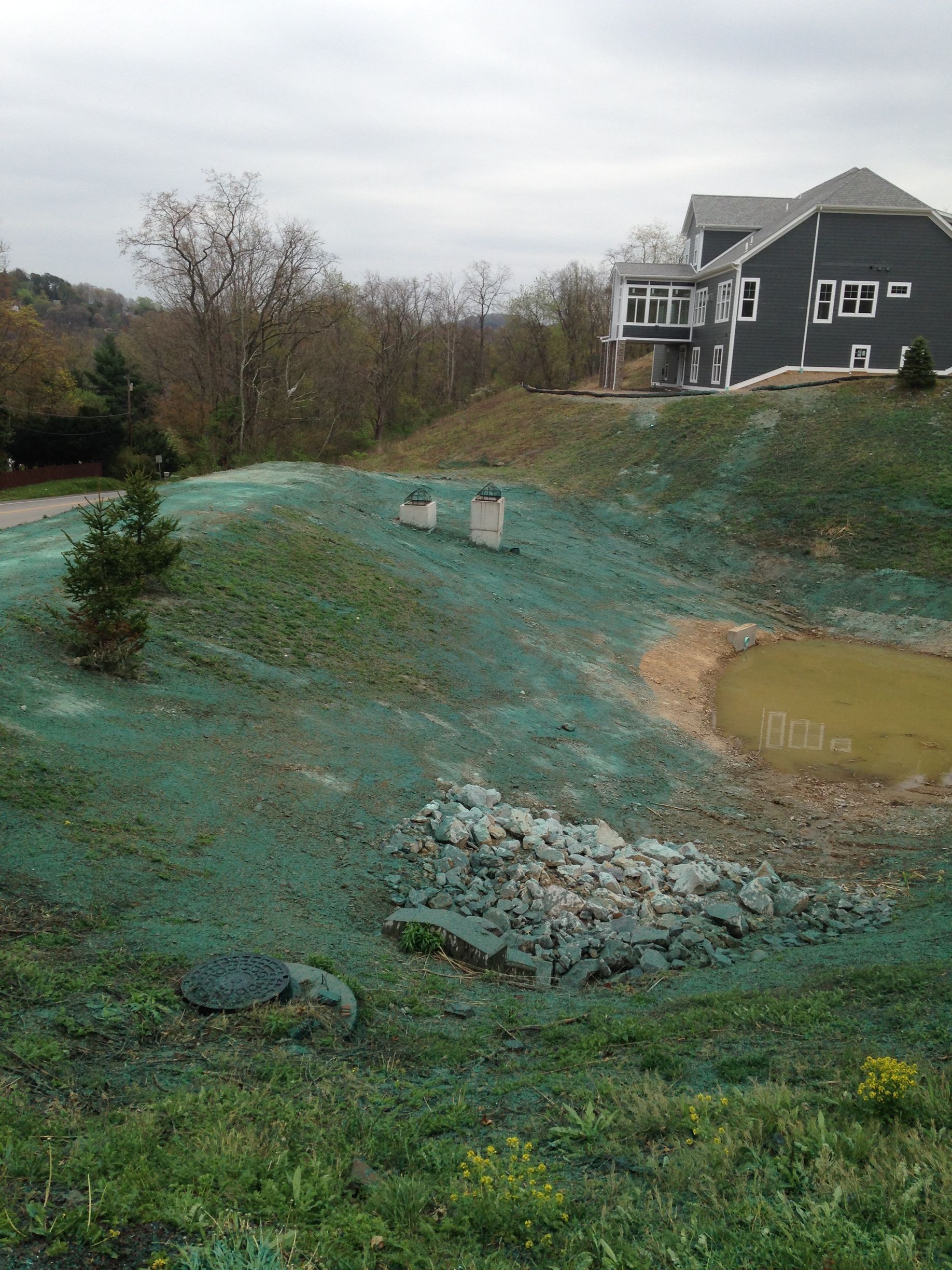 Construction site with green erosion blanket, muddy pond, and house on a hillside under an overcast sky