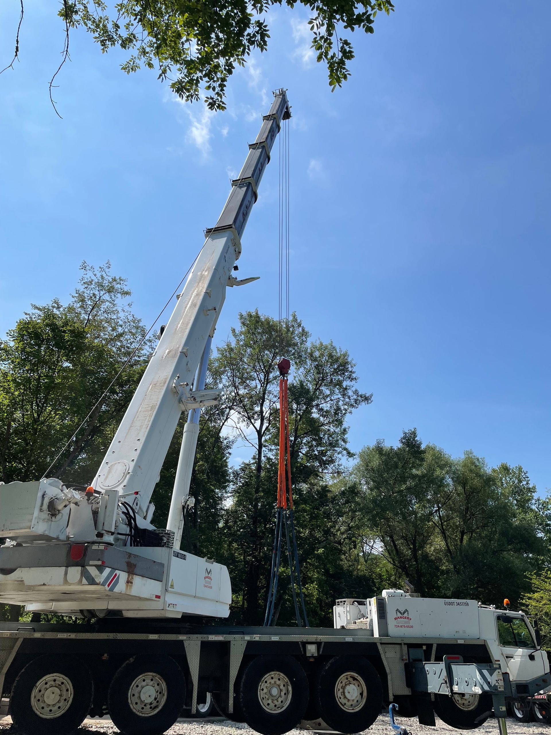 White crane truck lifting a red hanging load in a sunny, tree-lined area